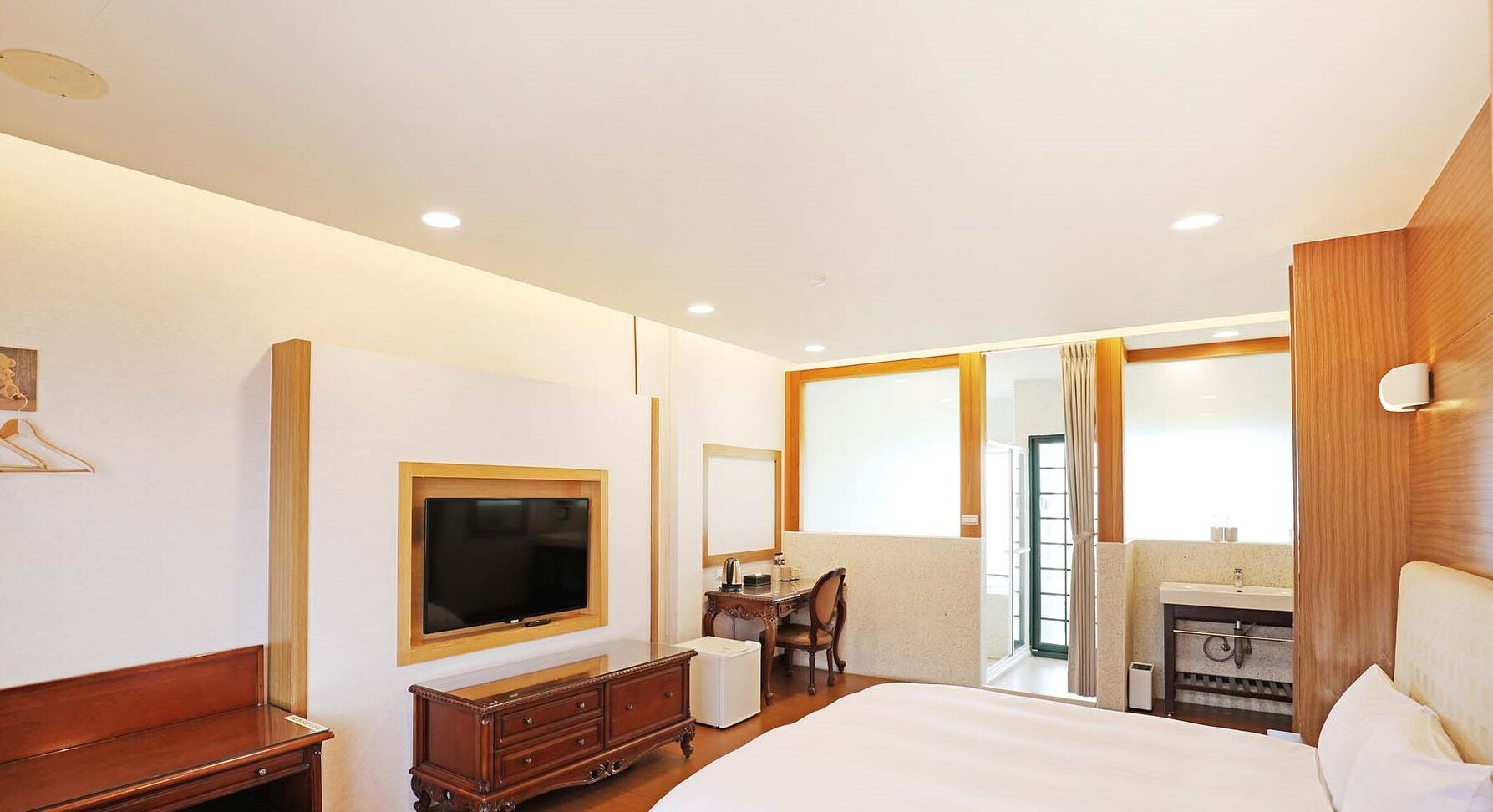 A hotel room with a large bed, a TV mounted on the wall, a desk with a chair, a small bathroom, and a window with curtains.