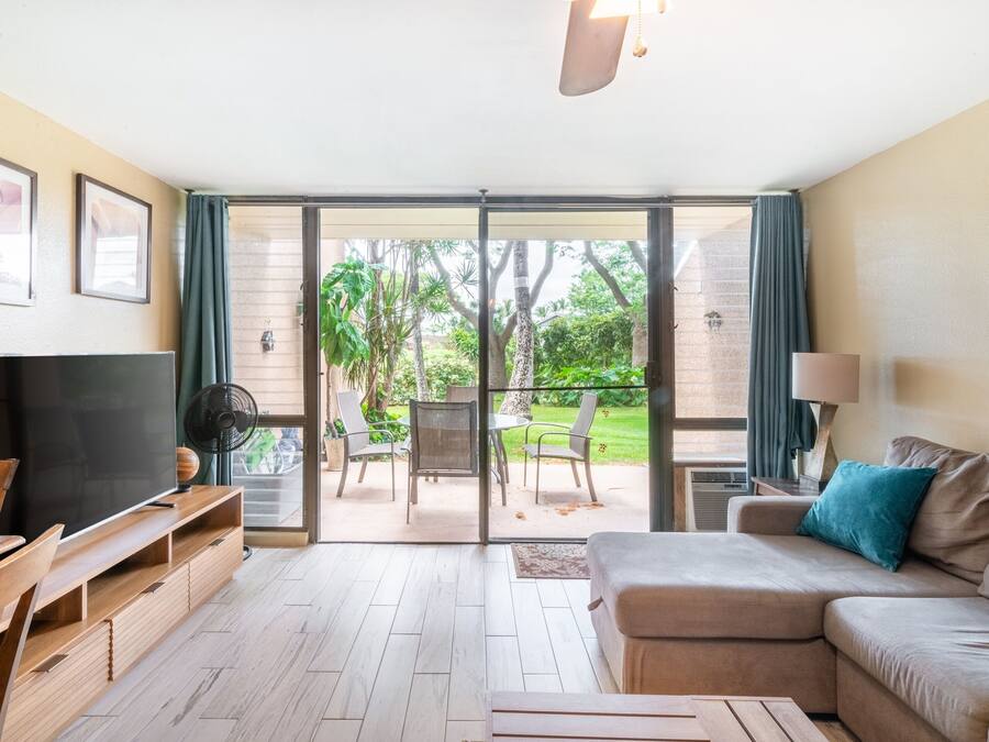 Una sala de estar con sofá, televisor, muebles de madera y un ventanal corredizo que da a un patio exterior con sillas y una mesa.