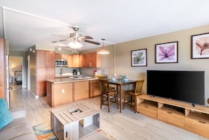Un appartement moderne comprenant une cuisine, un coin repas et un salon. Il est doté d’armoires en bois, d’un ventilateur de plafond, d’un téléviseur à écran plat et d’œuvres d’art accrochées aux murs.