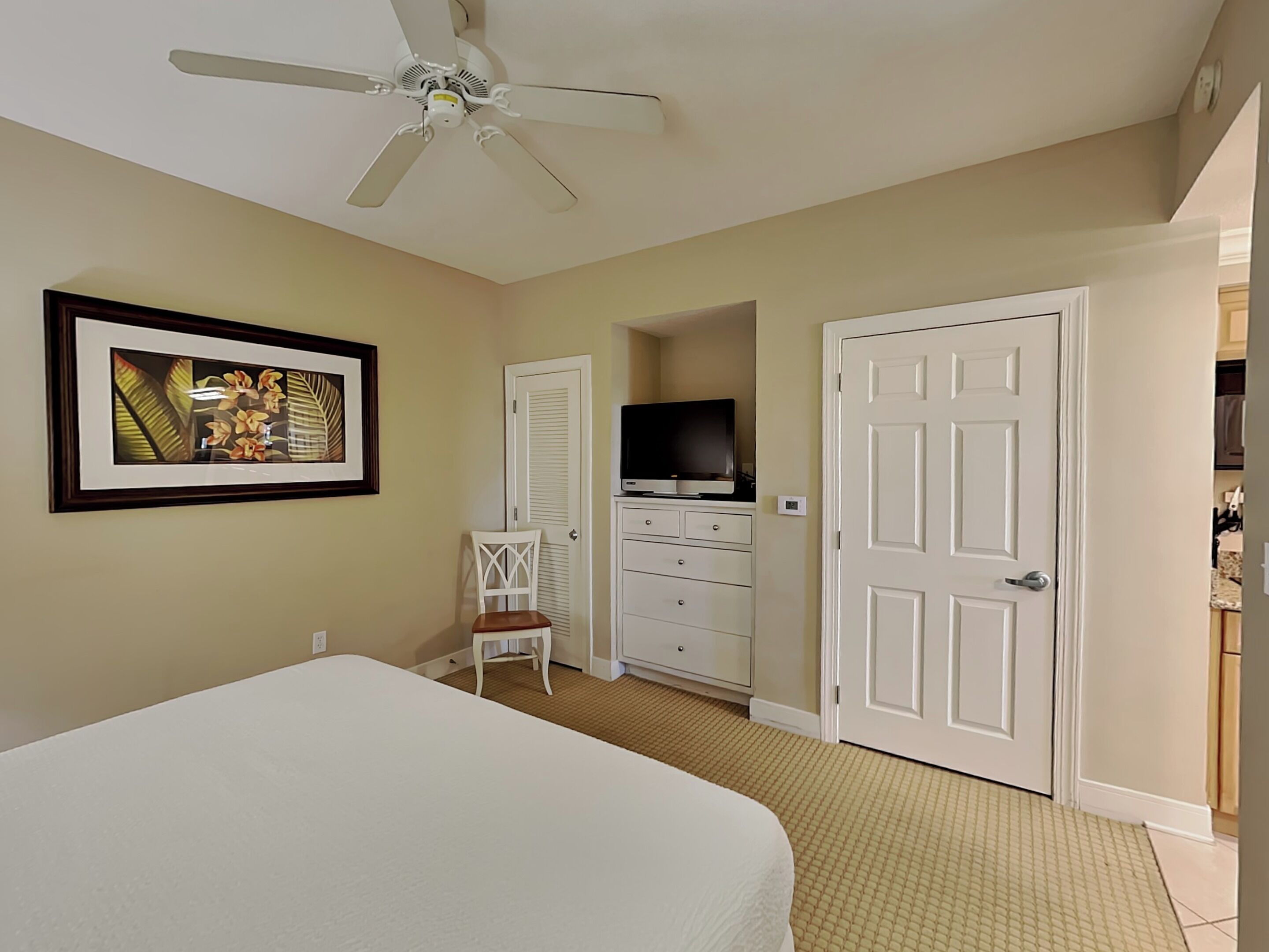 A bedroom with a bed, a ceiling fan, a TV, a dresser, and a door leading to another room.