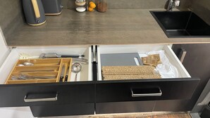 Une cuisine avec un plan de travail foncé, une machine à café, des bocaux d’épices et un évier. En dessous, on trouve des tiroirs ouverts contenant des ustensiles et une planche à découper.