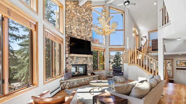 A spacious living room with a stone fireplace, large windows, a wooden staircase, and a cozy seating area.