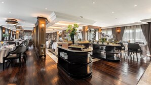 A spacious hotel dining area with multiple tables, a central bar, and a view of the outdoors. - Ambassador Signature Cruise - Lan Ha Bay (Bai Chay)