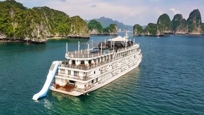 A large cruise ship with multiple decks and a water slide, surrounded by lush green hills and clear blue waters. - Ambassador Signature Cruise - Lan Ha Bay (Bai Chay)