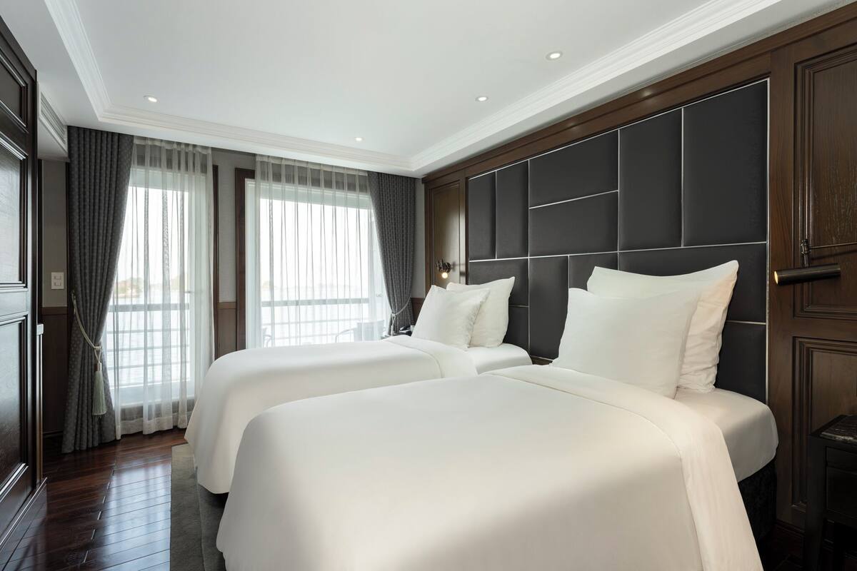 A hotel room with two beds, a large window with curtains, a dark headboard, and a wooden floor.