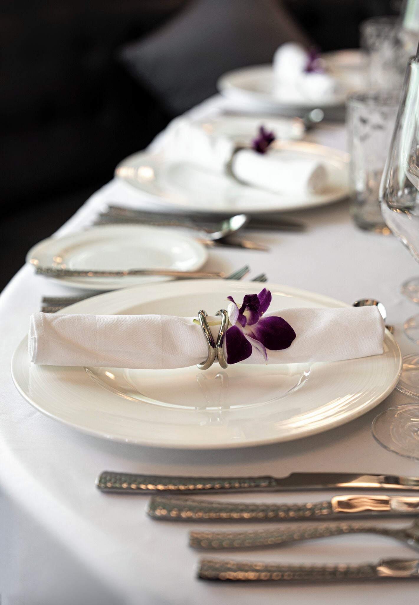 A neatly set dining table with white plates, silverware, and a folded napkin with a purple flower.