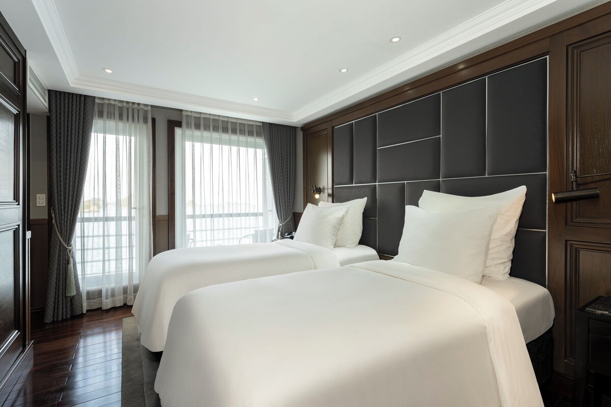 A hotel room with two beds, a large window with curtains, a dark headboard, and a wooden floor.