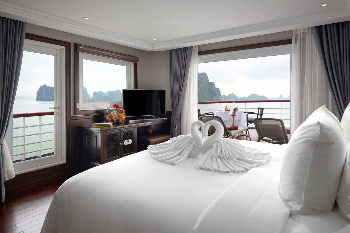 A hotel room with a bed, a TV, a dining table with chairs, and a view of the sea.