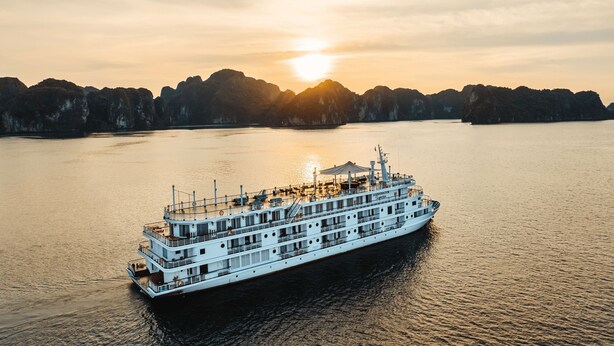 A large white river cruise ship with multiple decks and a covered upper deck, sailing on calm waters with a mountainous coastline in the background. - Ambassador Signature Cruise - Lan Ha Bay (Bai Chay)