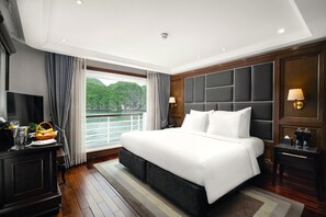 A spacious hotel room with a large bed, a television, a desk with a lamp, and a view of a body of water. - Ambassador Signature Cruise - Lan Ha Bay (Bai Chay)