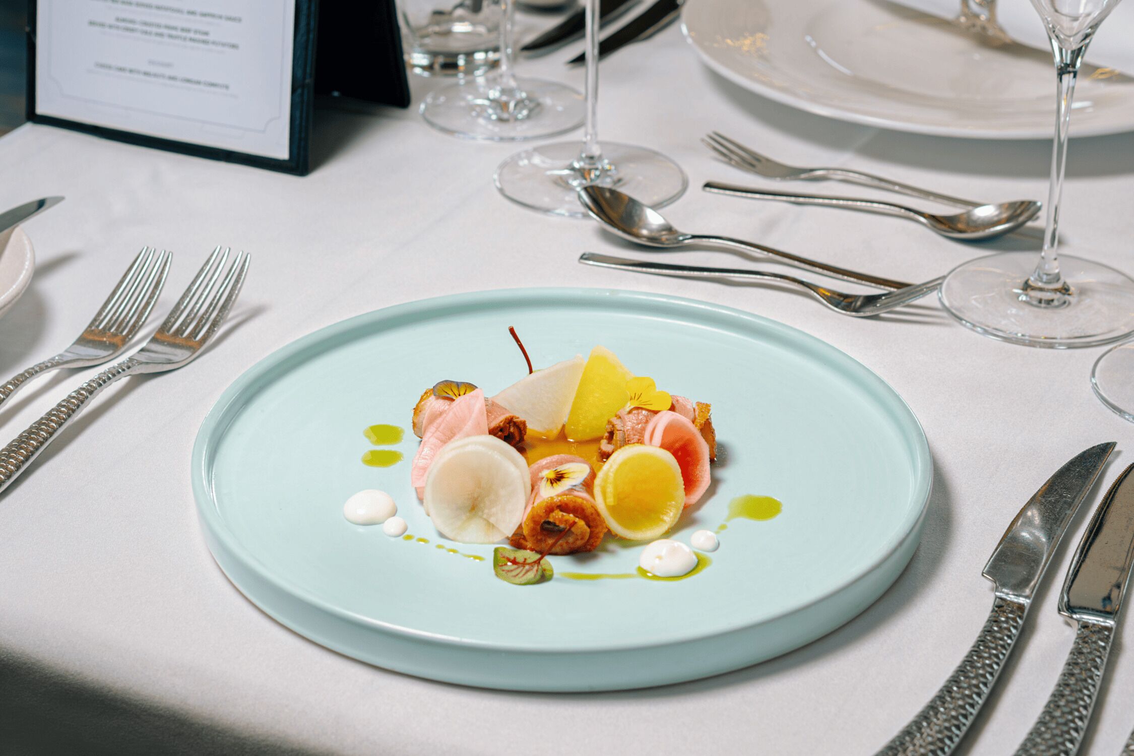 A formal dining table with a light blue plate of food, silverware, and wine glasses.