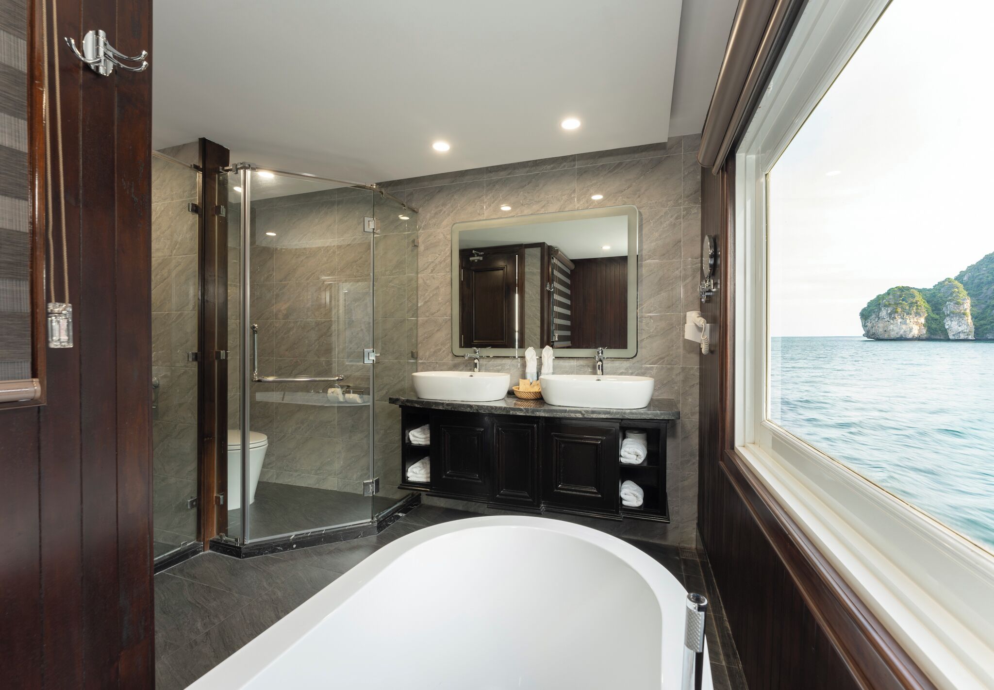 Ein modernes Badezimmer mit einer gläsernen Duschkabine, zwei Waschbecken, einer großen Badewanne und Meerblick durch ein Fenster.