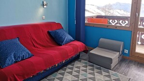 Un sofá rojo con cojines azules, un sillón gris y una alfombra estampada en una habitación con paredes azules y vistas a las montañas.