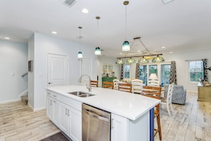 A modern kitchen with white cabinetry, a stainless steel dishwasher, and a large island with a sink. There are hanging pendant lights above the island and a dining area with wooden chairs and a patterned sofa.
