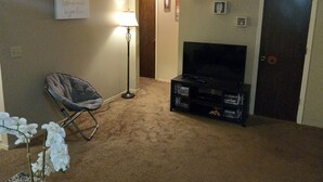 Un salon avec un téléviseur à écran plat posé sur un meuble, une lampe sur pied, un fauteuil et une petite table sur laquelle se trouve une plante.