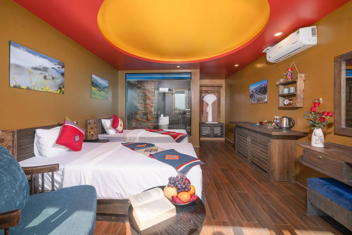 A hotel room with two beds, a bathroom with a glass shower, a wooden desk, and a small kitchen area.