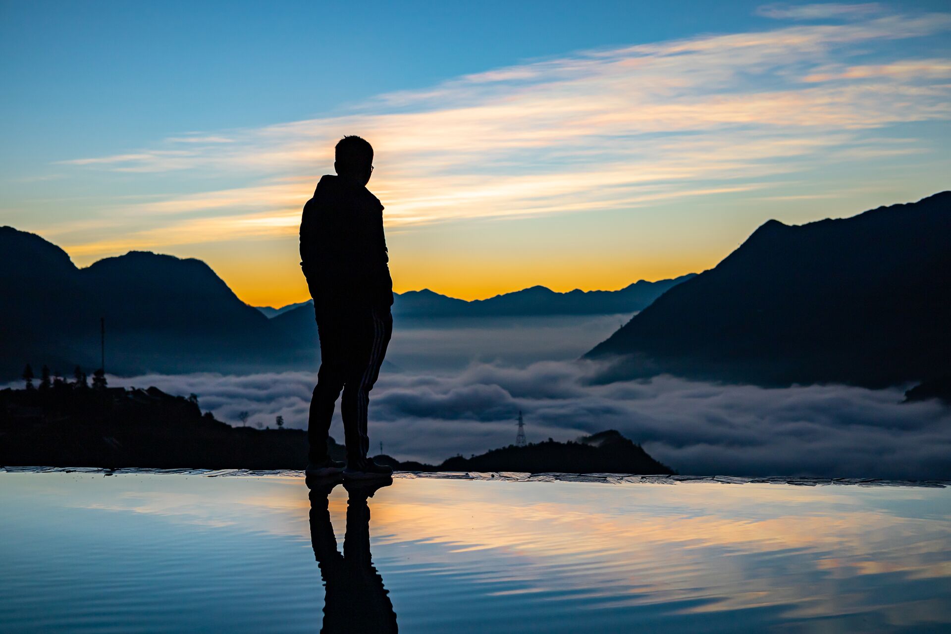 A person standing on a reflective surface with a mountainous landscape and clouds in the background.