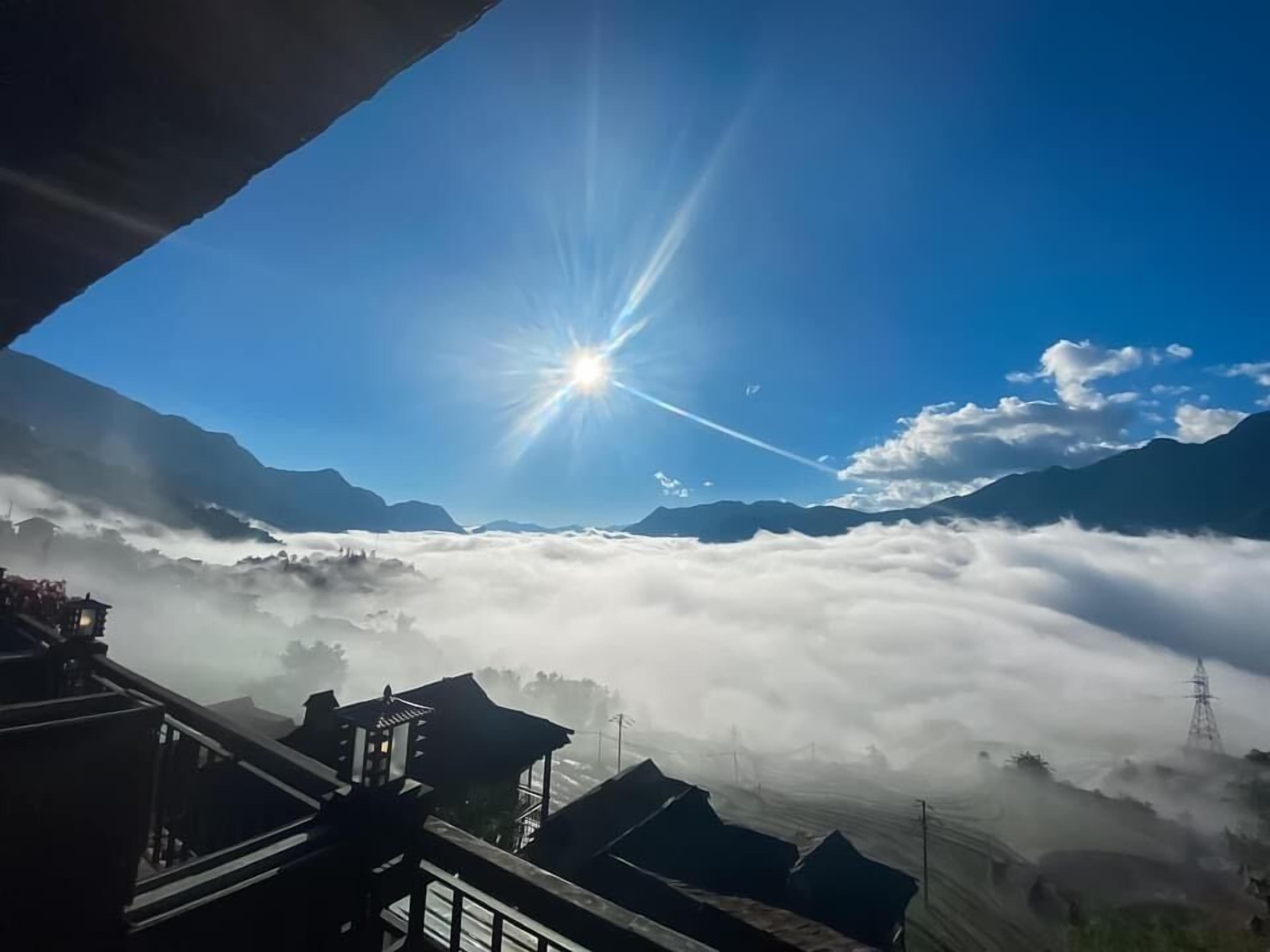 Un paisaje montañoso con el sol brillando entre las nubes, cielo despejado y vista de un valle con edificios.