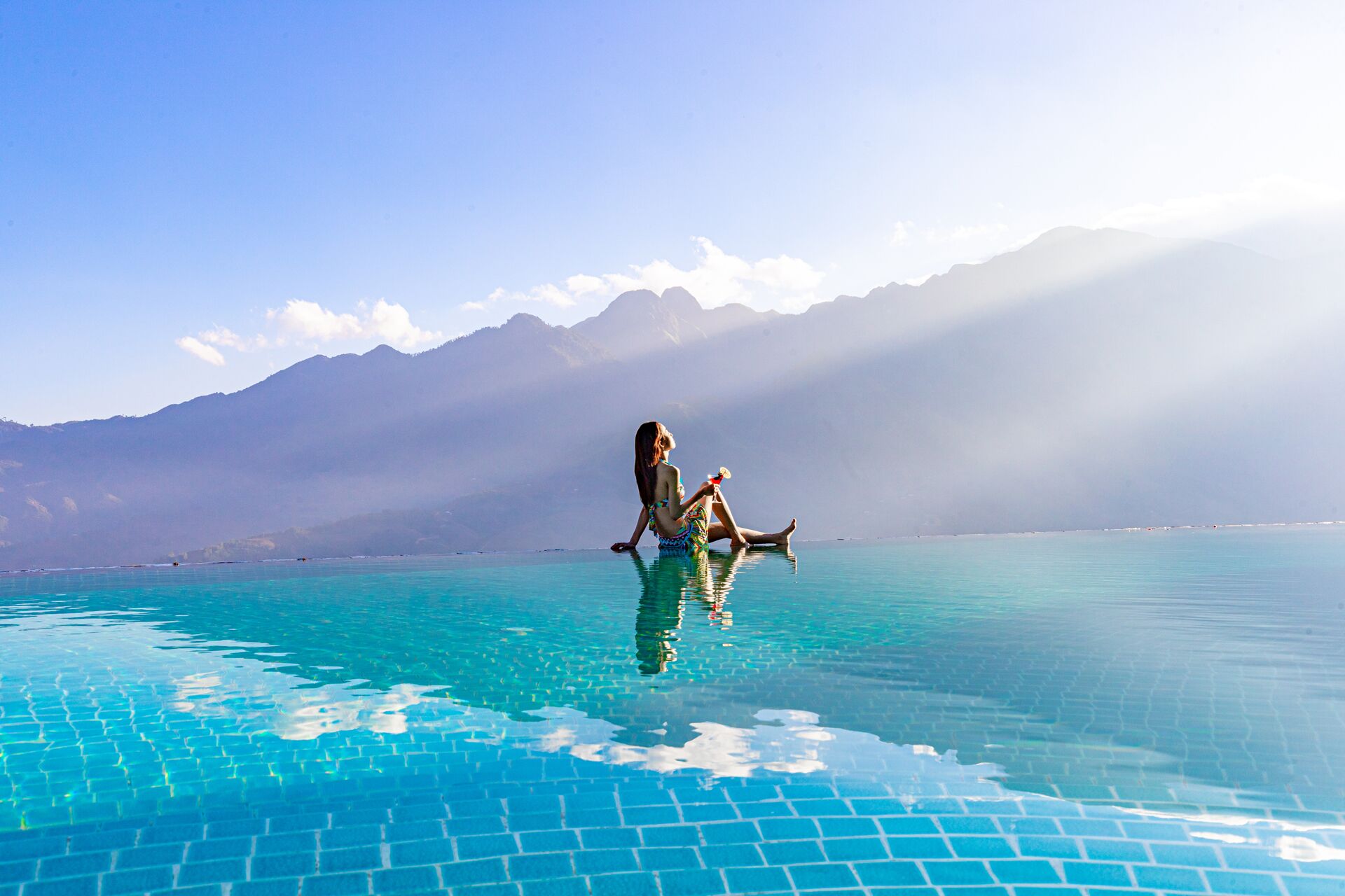 Une personne assise au bord d’une piscine à débordement aux eaux d’un bleu limpide, avec une chaîne de montagnes en arrière-plan.