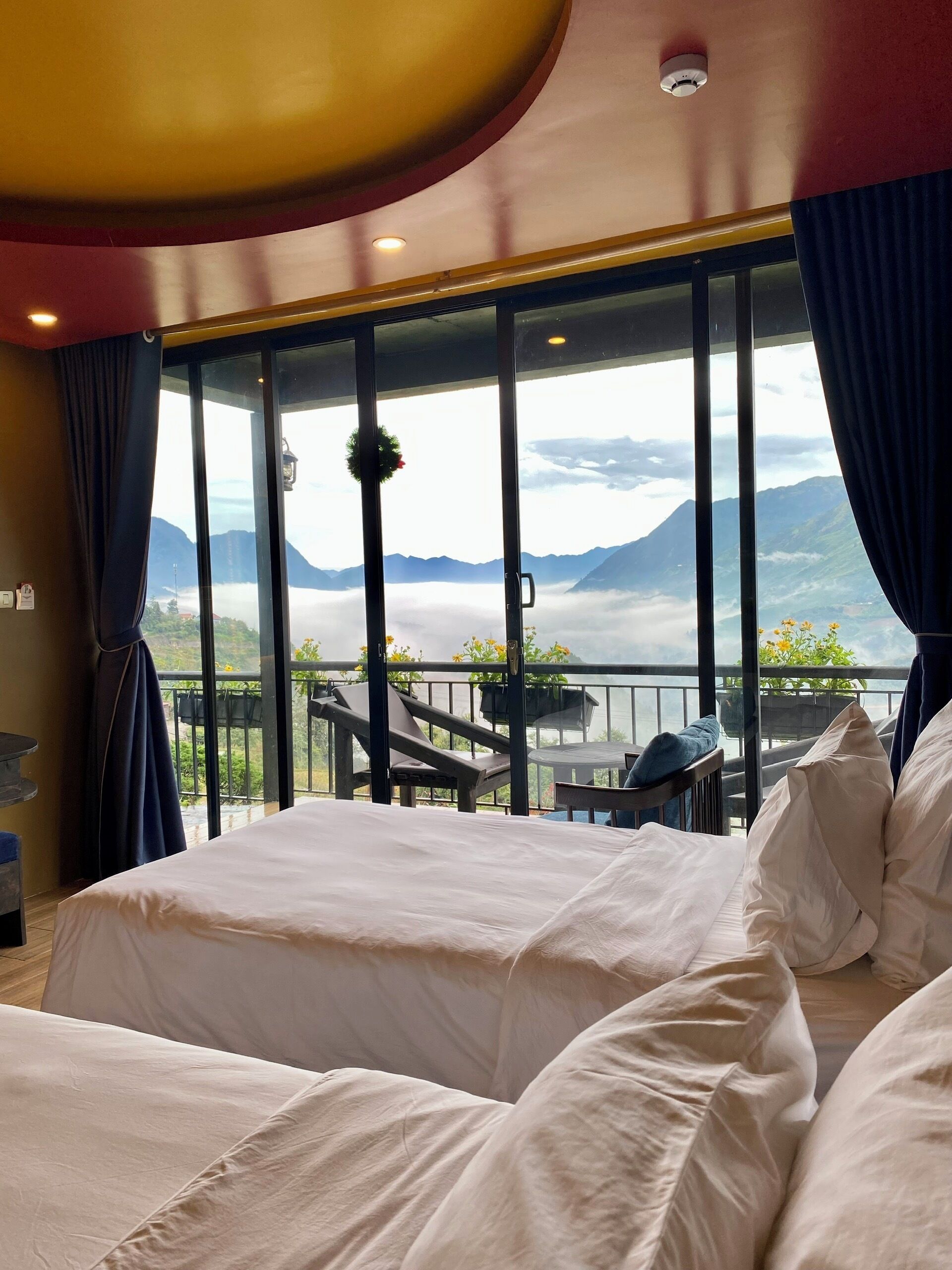 Une chambre d’hôtel avec un grand lit, un balcon offrant une vue sur les montagnes et les nuages, et un plafond orné d’un luminaire circulaire.