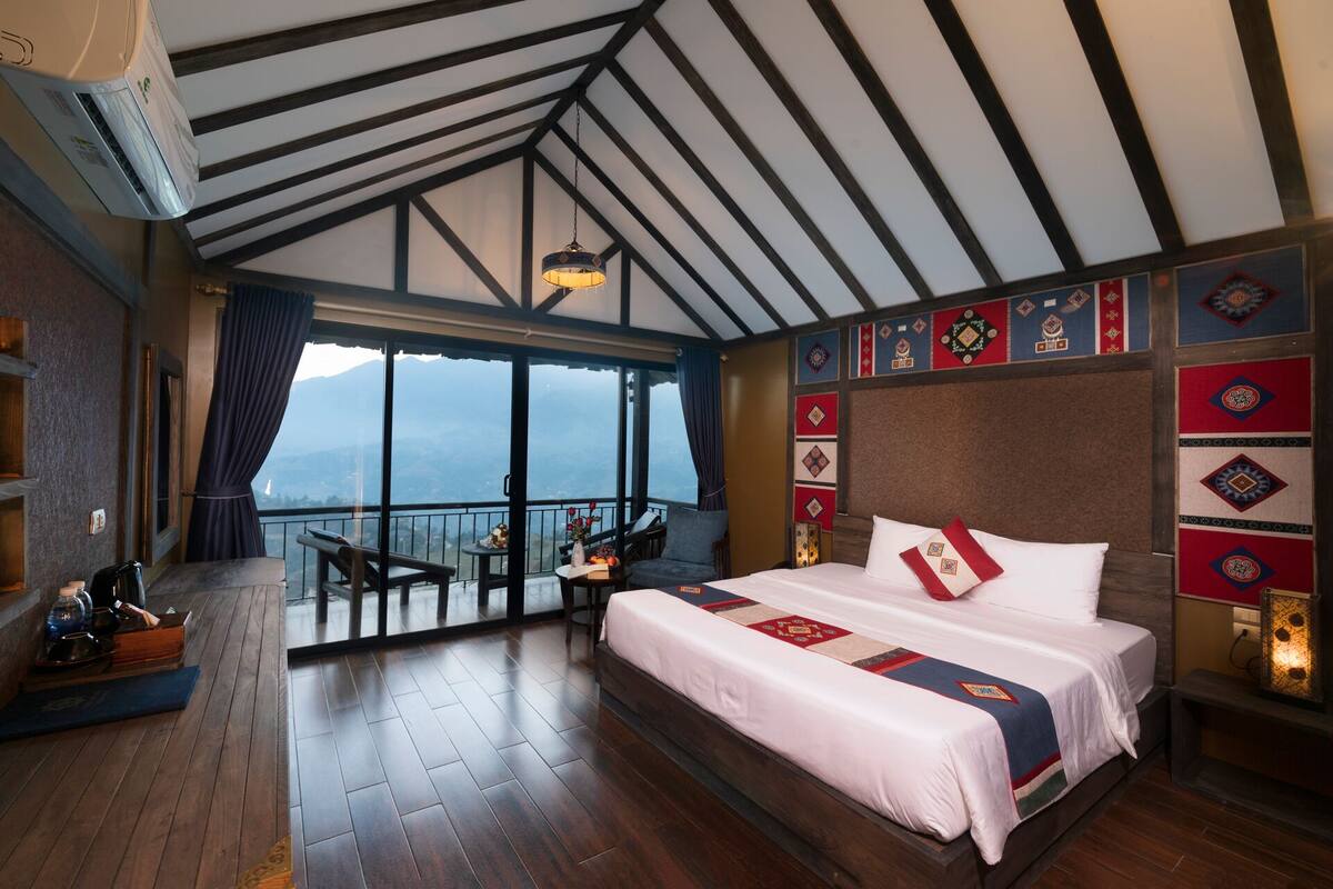 A hotel room with a large bed, a balcony with a view, and a wooden floor.