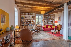 Una sala de estar acogedora con techo de madera, un sofá rojo, un sillón beige y una estantería llena de libros y objetos decorativos.