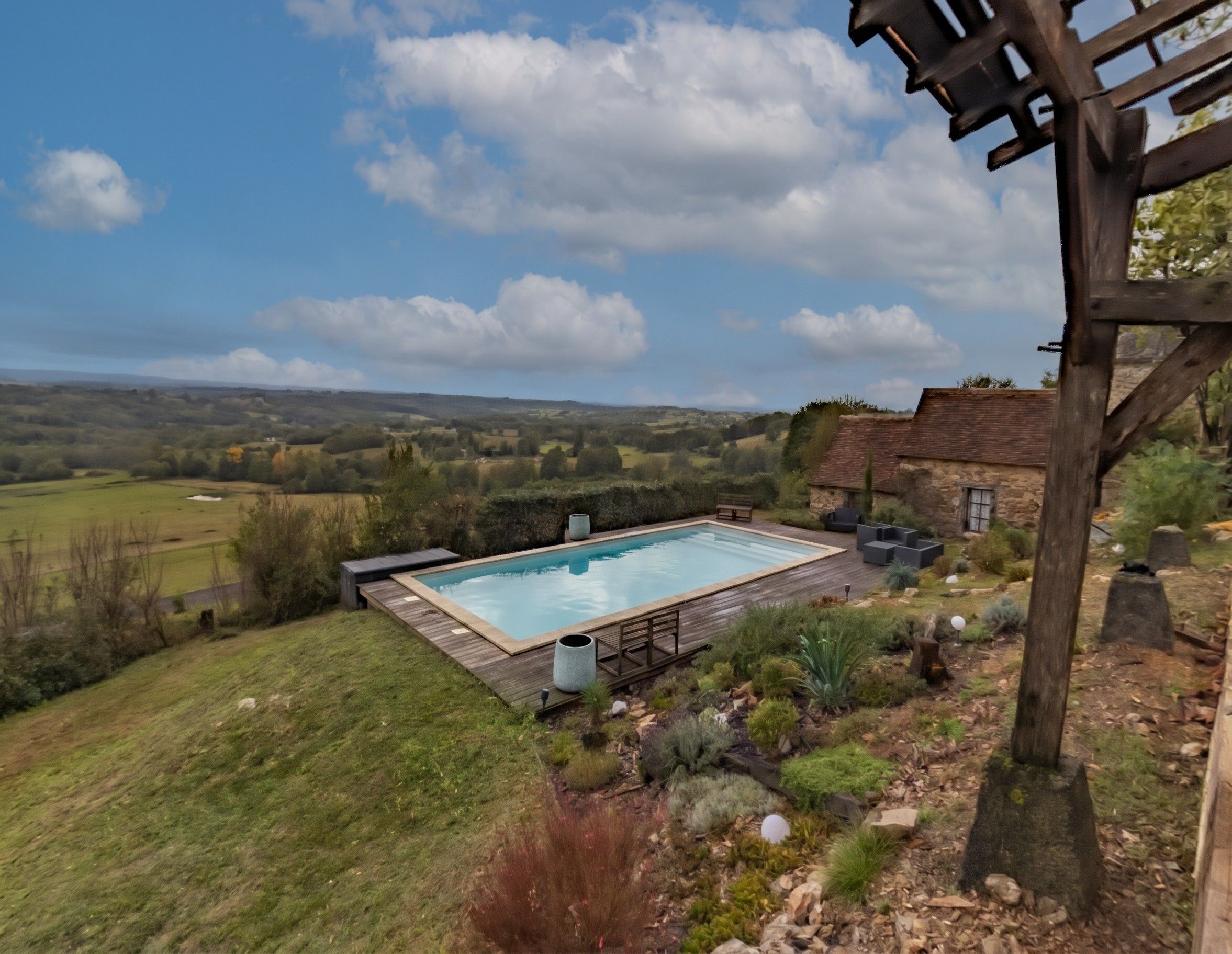 Une piscine rectangulaire dotée d’une terrasse en bois, entourée d’un jardin aménagé avec différentes plantes et d’un mur en pierre.
