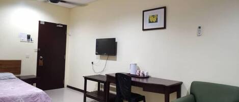 A hotel room with a bed, a desk with a chair, a green armchair, a flat-screen TV, and an air conditioning unit.