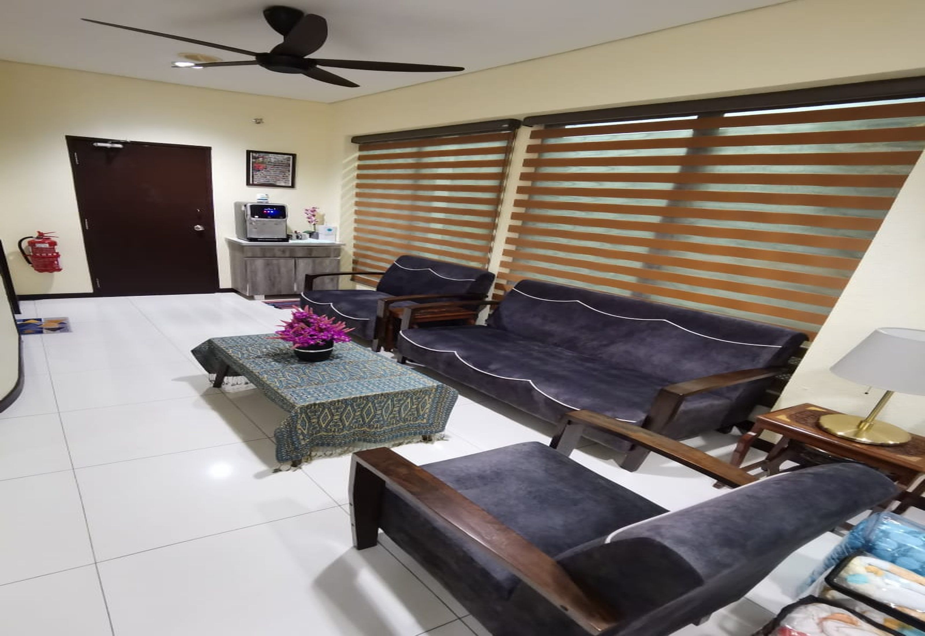 Una sala de estar con un sofá, dos sillones, una mesa de centro y un televisor. Se observa un ventilador de techo, un extintor y una ventana con persianas.