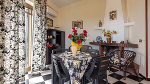 Ein Essbereich mit Schachbrettmuster-Boden, einem runden Tisch mit floralem Tischläufer, schwarzen Stühlen, einer Vase mit roten Blumen, einem Kamin und einem Bücherregal.