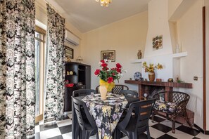 A dining area with a checkered floor, a round table with a floral tablecloth, black chairs, a vase with red flowers, a fireplace, and a bookshelf. - Vacation Home "Zina" with Sea View, 2 Private Terraces & 2 Balconies (Altavilla milicia)