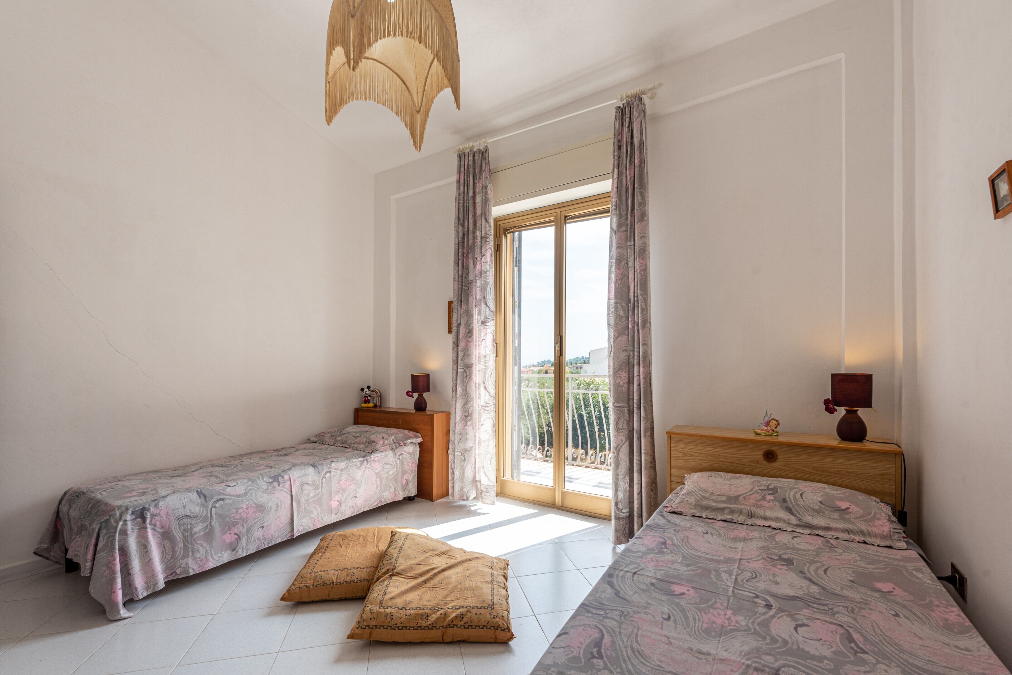 Une chambre avec deux lits, une grande fenêtre avec des rideaux, une tête de lit en bois et un luminaire suspendu.