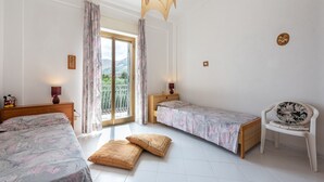 Ein Schlafzimmer mit zwei Betten, einem Sessel, einem Fenster mit Vorhängen, einem Balkon und einer Pendelleuchte.