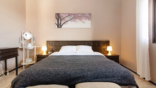 A hotel room with a bed, bedside tables, lamps, a clock, a painting, and a mirror.