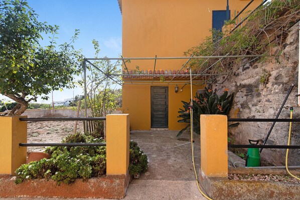 A yellow building with a door, a small garden with plants, and a hose. - Vacation Rental 'Apartamento Brownie' with Sea View, Private Terrace and Wi-Fi (Firgas)