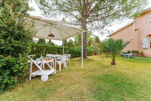 A covered outdoor seating area with tables and chairs, surrounded by greenery and a building in the background.