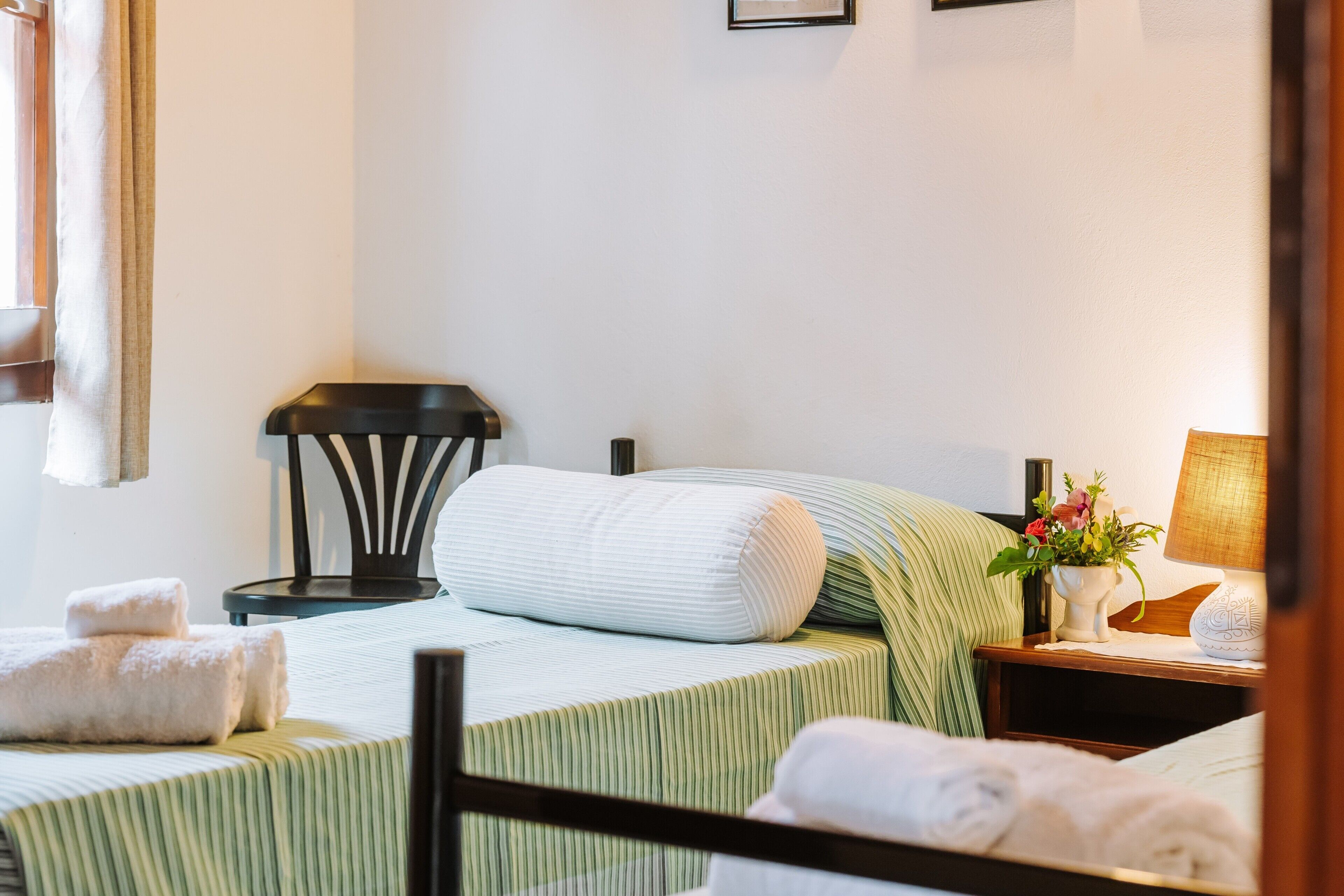 Een hotelkamer met twee bedden, een nachtkastje met een lamp, een stoel en een raam met gordijnen.