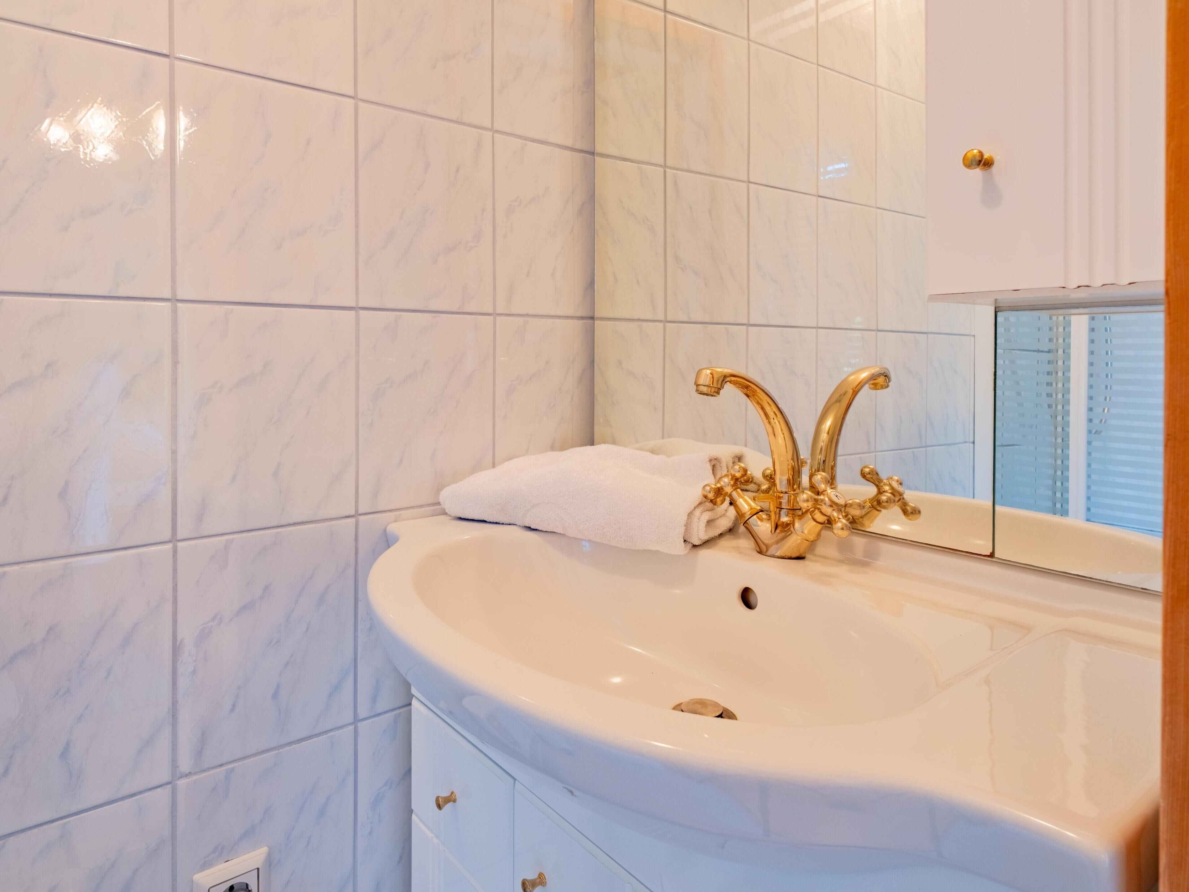 Een badkamer met een wit marmeren wastafel, gouden kraanwerk en een spiegel die een raam met luiken weerspiegelt.