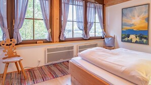 Een slaapkamer met een houten bed, een radiator, een raam met gordijnen, een stoel en een schilderij aan de muur.