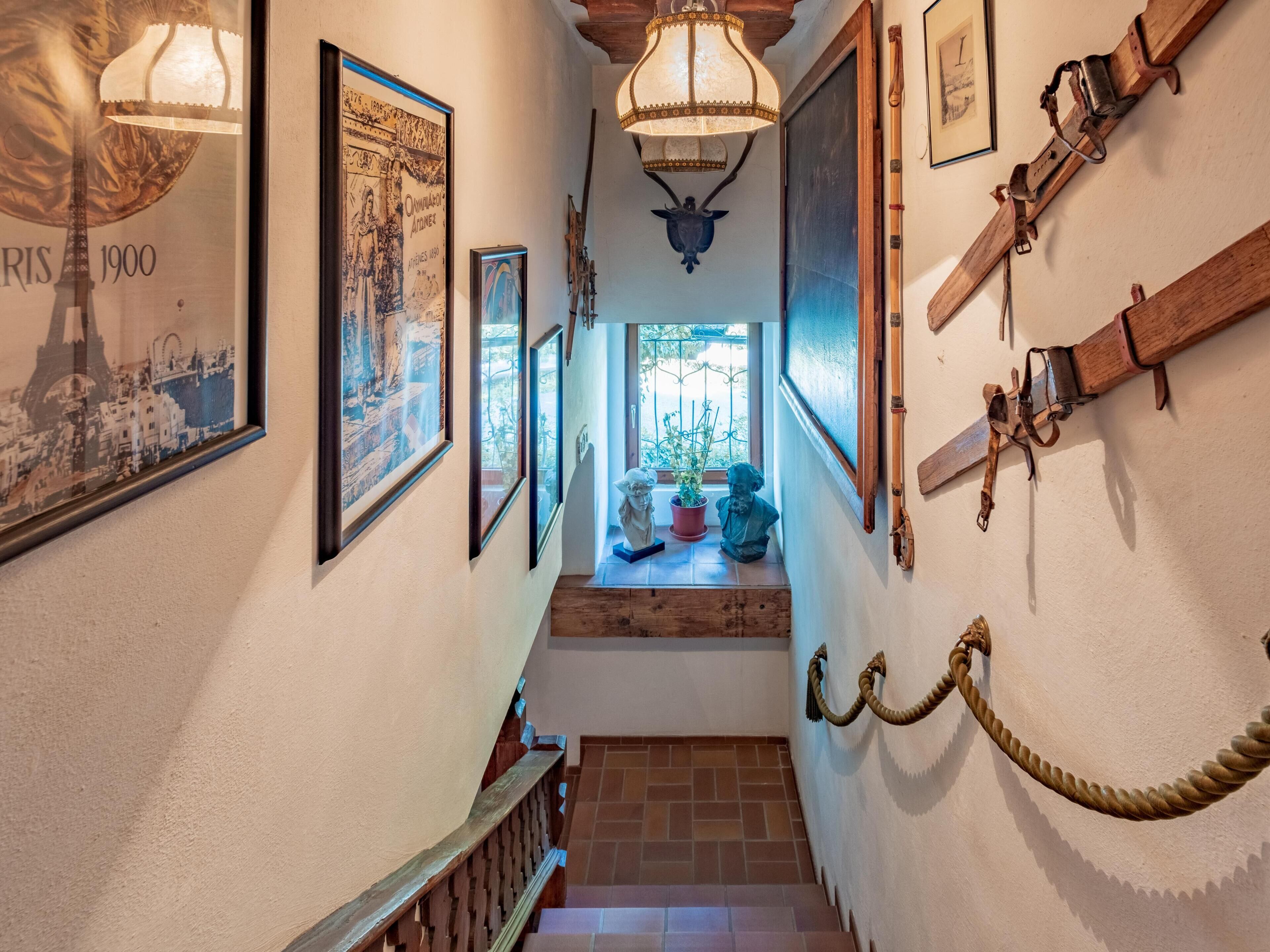 Un couloir étroit orné de tableaux encadrés, un escalier en bois et une fenêtre avec des plantes.