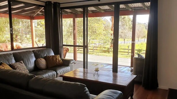 A spacious living area with a leather sofa, a wooden coffee table, and large windows offering a view of a garden and street. - Blacksmith Corner (Nannup)