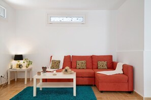 Ein Wohnzimmer mit rotem Sofa, weißem Couchtisch, blaugrünem Teppich und einem kleinen Beistelltisch mit Lampe.