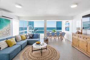 A modern living room with a sofa, coffee table, and dining area, featuring large windows with ocean views. - Absolute Oceanfront Yamba - oceanstays (Yamba)