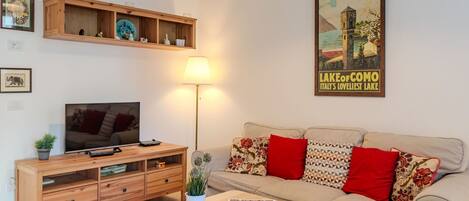 A cozy living room with a TV, wooden entertainment unit, sofa with patterned cushions, and a poster of Lake Como.