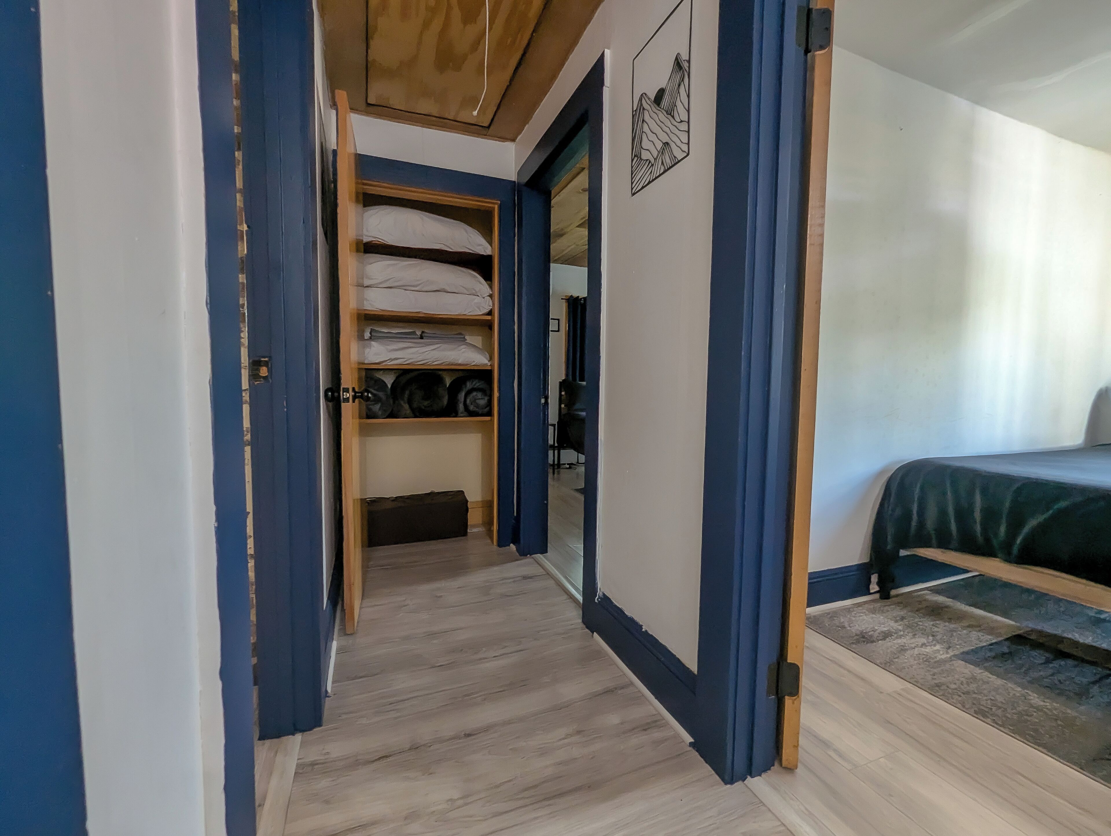 A narrow hallway with a wooden ceiling, a bed with a green bedspread, and a wardrobe with shelves containing folded items and a suitcase.
