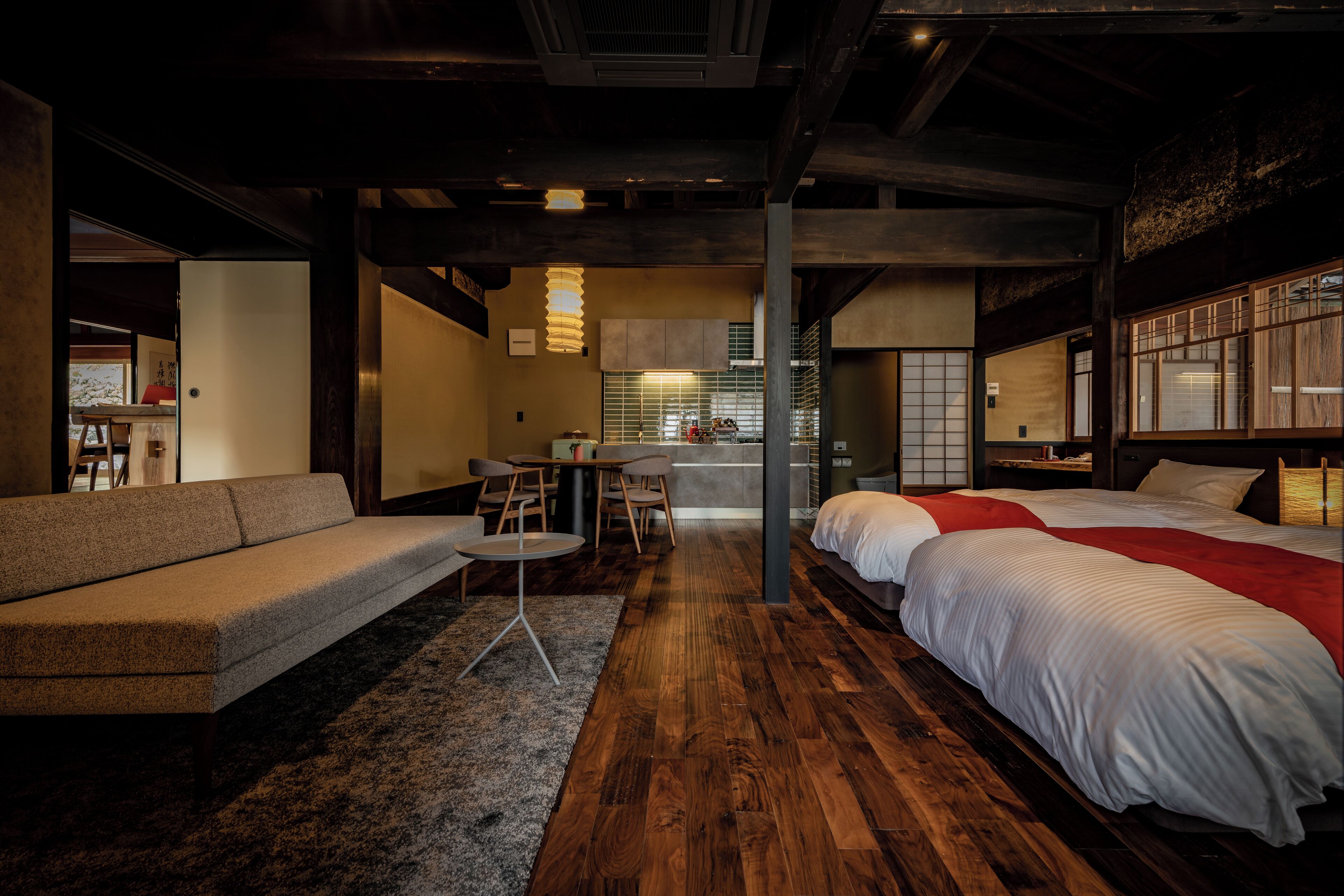 A modern hotel room with a wooden floor, a sofa, a bed with a red blanket, a small table with chairs, and a kitchen area in the background.