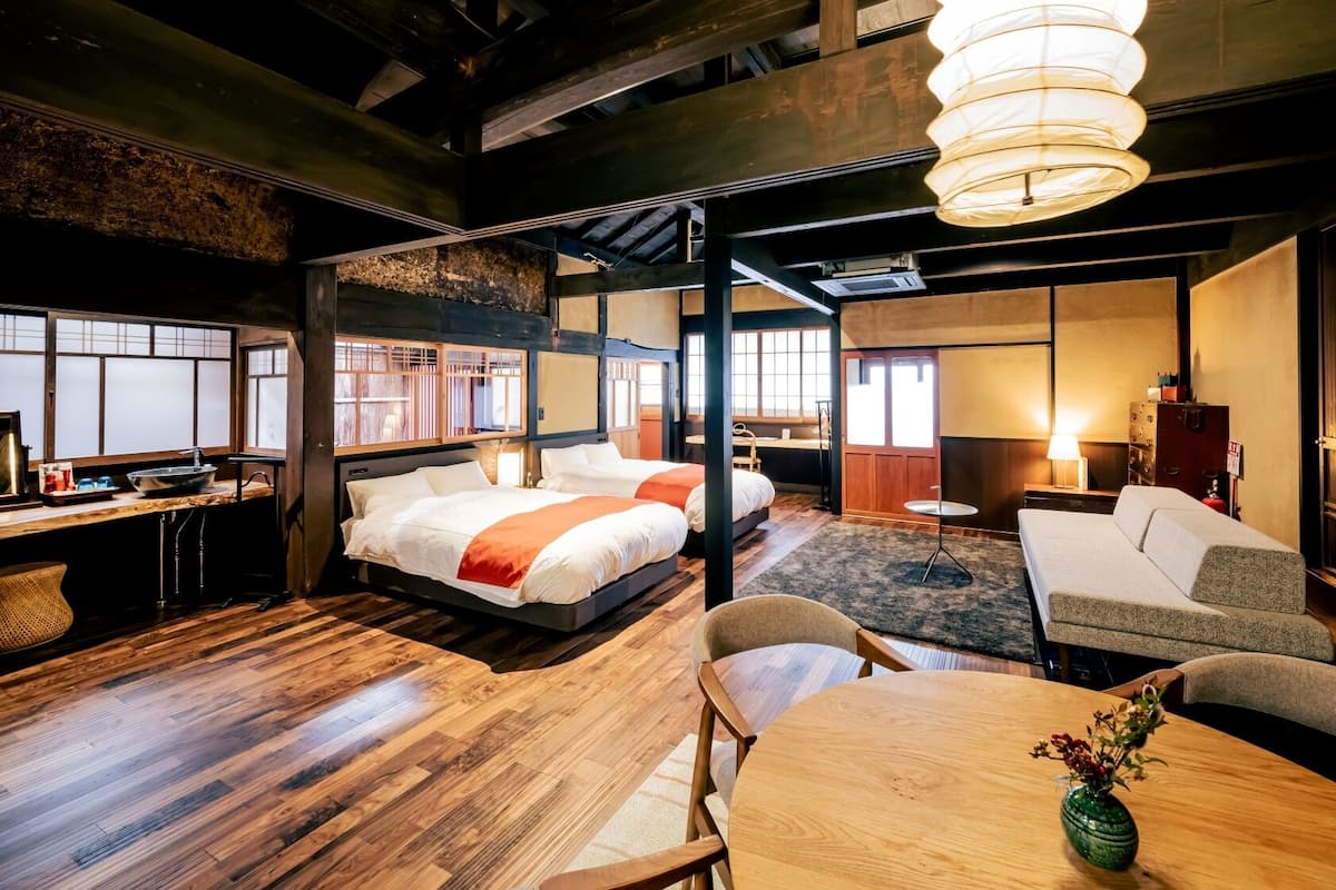 Une chambre d’hôtel avec un plancher en bois, deux lits, un bureau avec un ordinateur, un canapé et une table à manger agrémentée d’un vase.