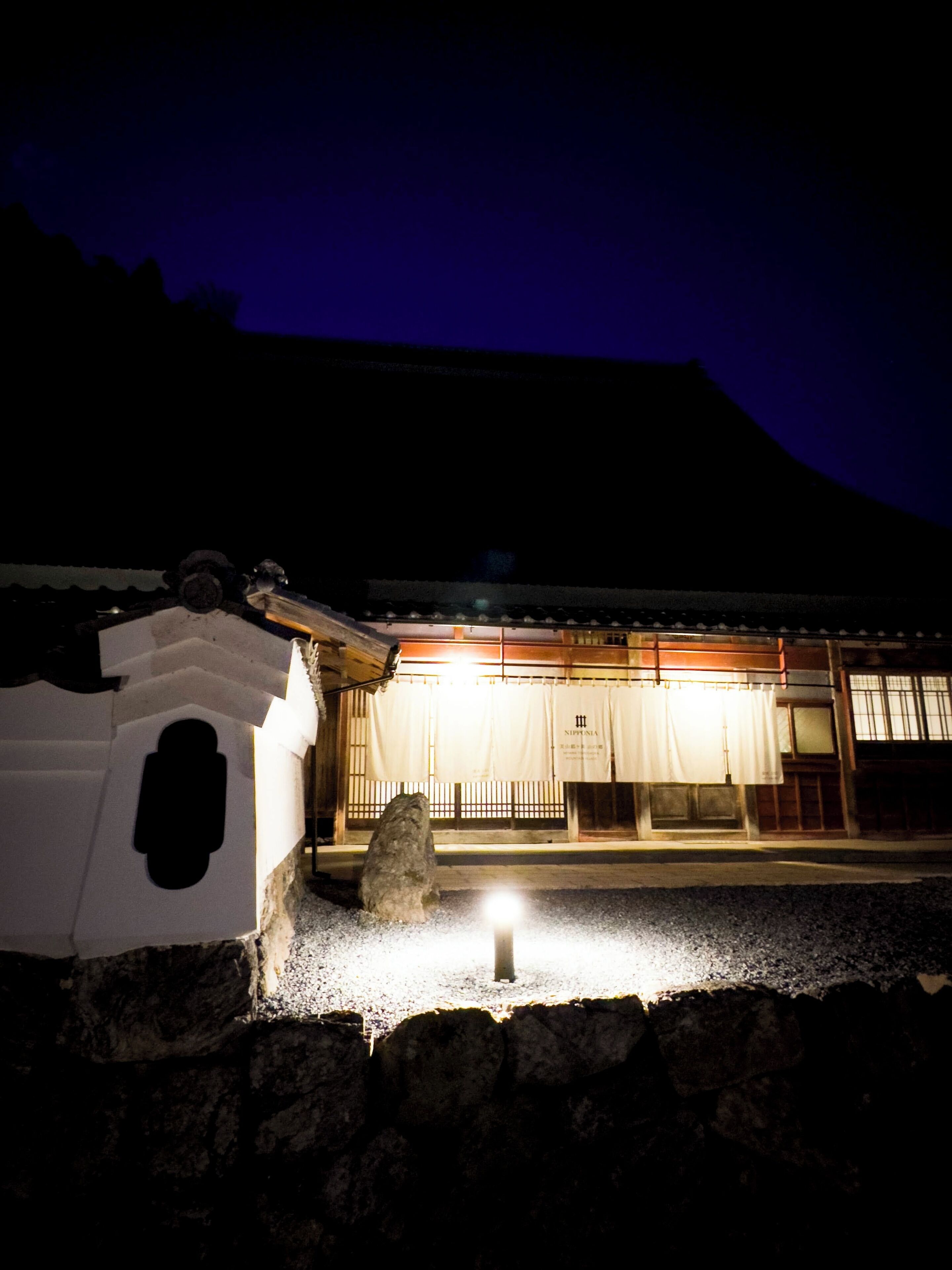 A traditional Japanese building with a stone lantern in the foreground, a wooden fence, and a sign with Japanese characters.