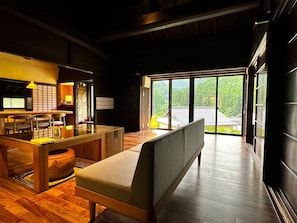 A modern interior with a wooden dining table, a cushioned bench, and a large window offering a view of a traditional house and greenery. - NIPPONIA Miyama Tsurugaoka Mountain Village (Nantan)