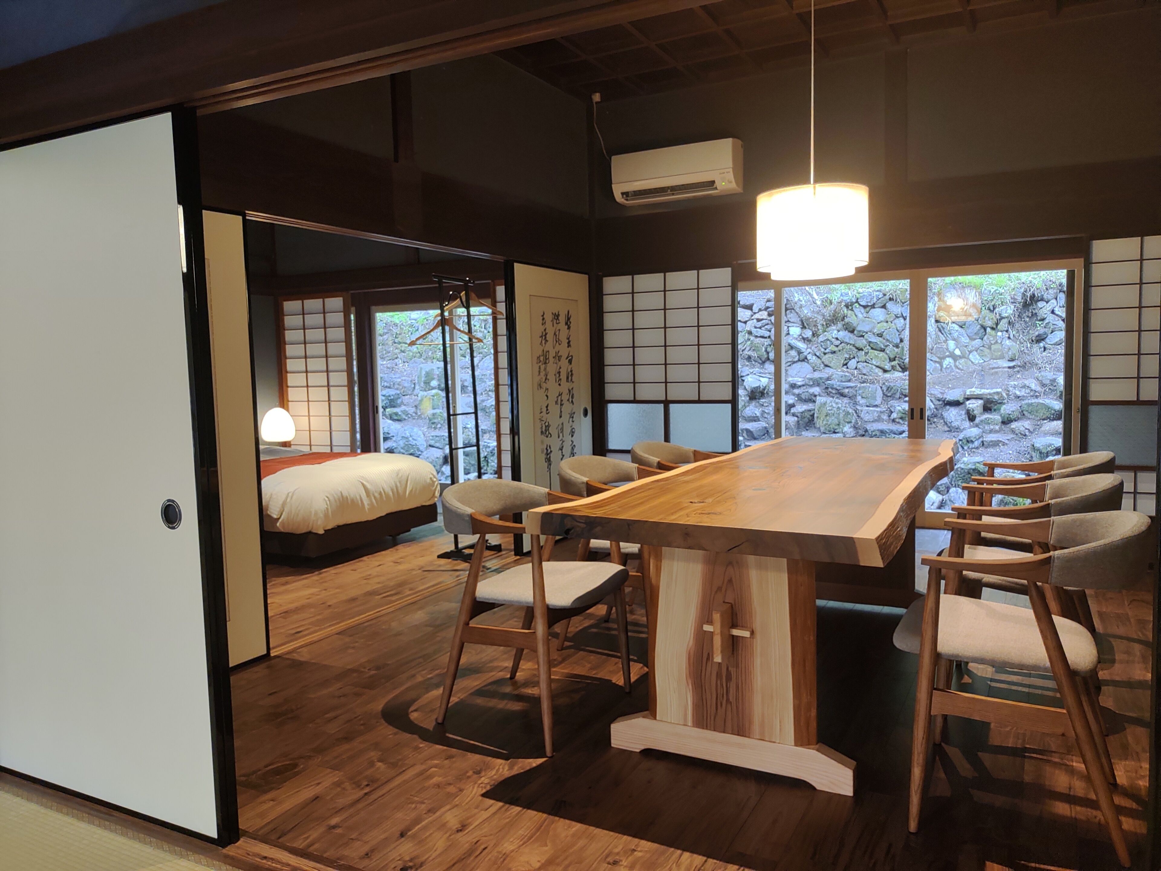 A traditional Japanese-style room with tatami flooring, a wooden dining table with chairs, a bed with a white comforter, and a view of a stone garden through large windows.